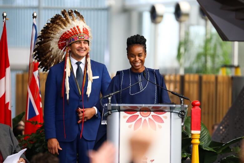 A person stands behind a podium next to a man wearing a headdress.