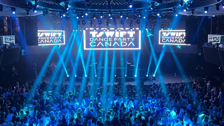 High wide shot of people dancing in a club, with the camera focused toward the stage. Screens in the room read T. Swift Dance Party.