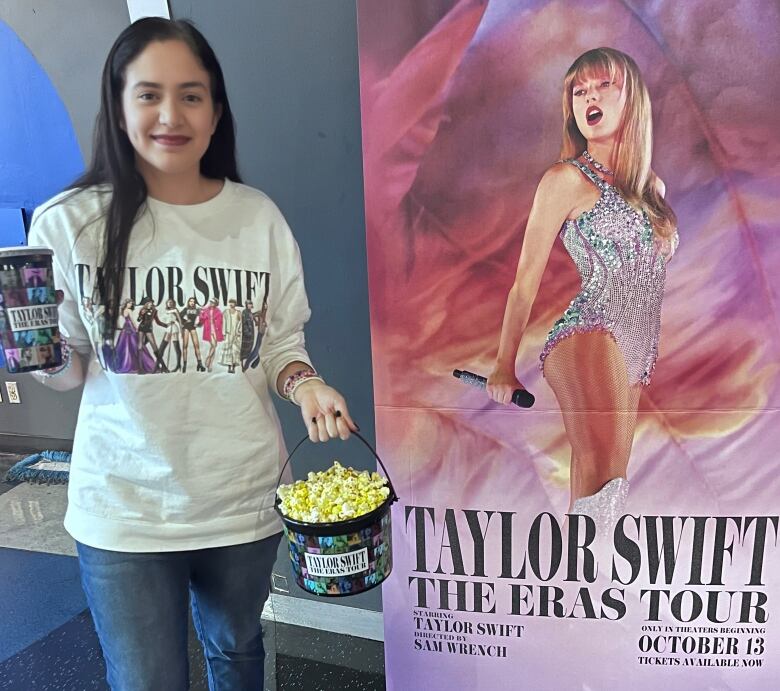 A woman with long hair, wearing a taylor swift t-shirt, holds a bucket of pop corn and drink cup, both with a commemorative Eras Tour print.