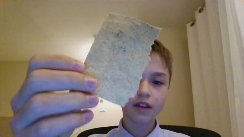 A teen boy with sandy blonde hair wearing a white dress shirt holds up a square of paper.