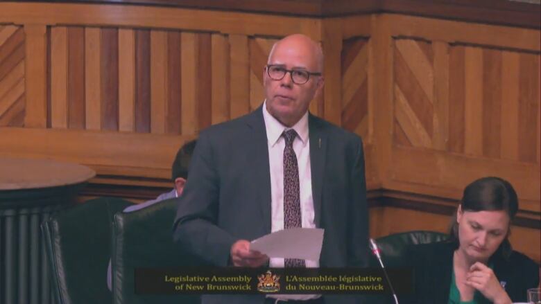 A bald man in a suit and tie is standing and speaking.