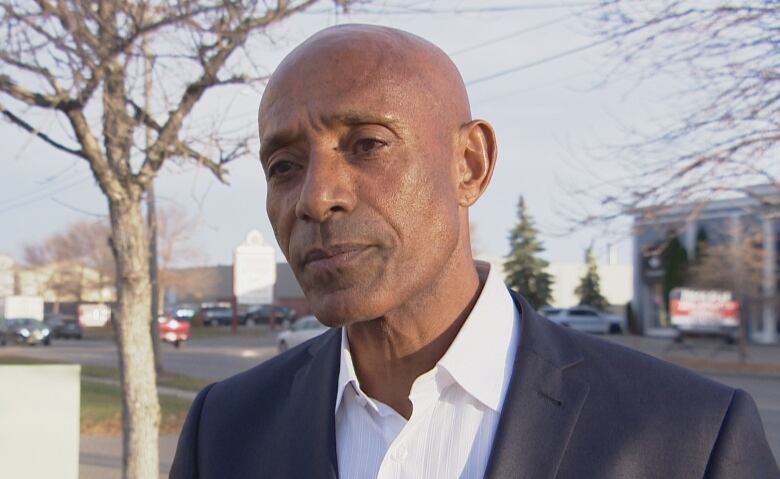 Bald man in suit stands outside in fall.