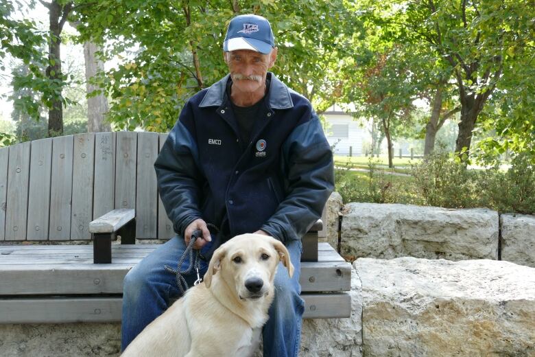 A man wearing a dark-coloured jacked is sitting on a bench in a park, holding his dog with a leash. 