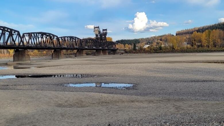 Low water levels are seen, with a gravel river bed with a few small puddles of water. In the background is a metal railway bridge and blue sky.