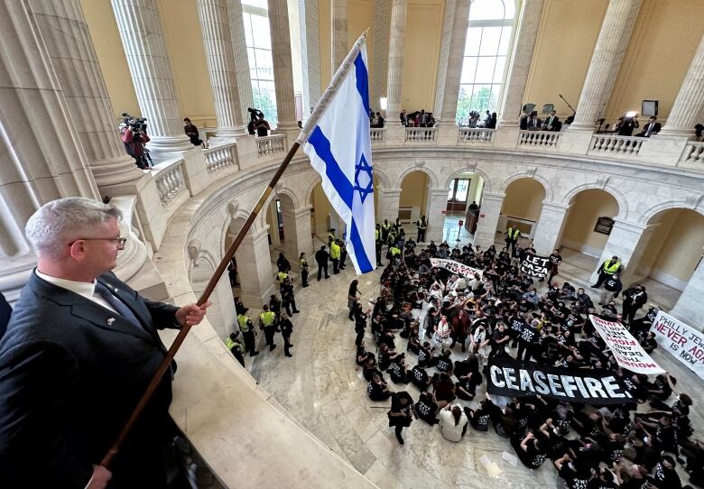 Man holds Israeli flag on second floor, above crowd of protesters on first floor of U.S. Capitol