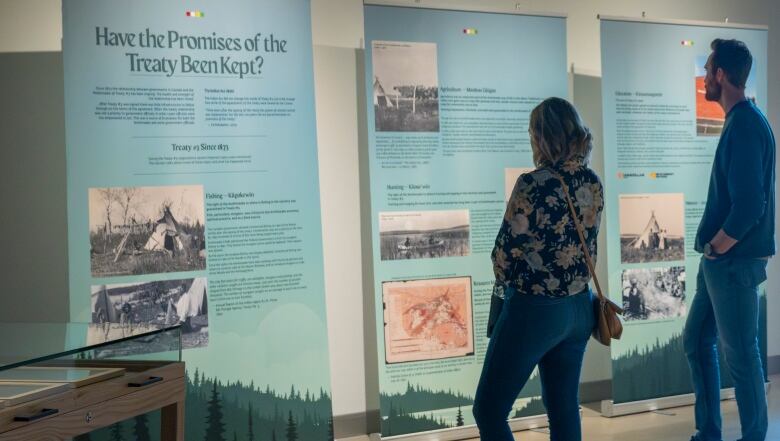 A man and woman are seen observing three panels that are hung on a wall, which include pictures and text. The headline of the first panel says 'Have the Promises of the Treaty Been Kept?'