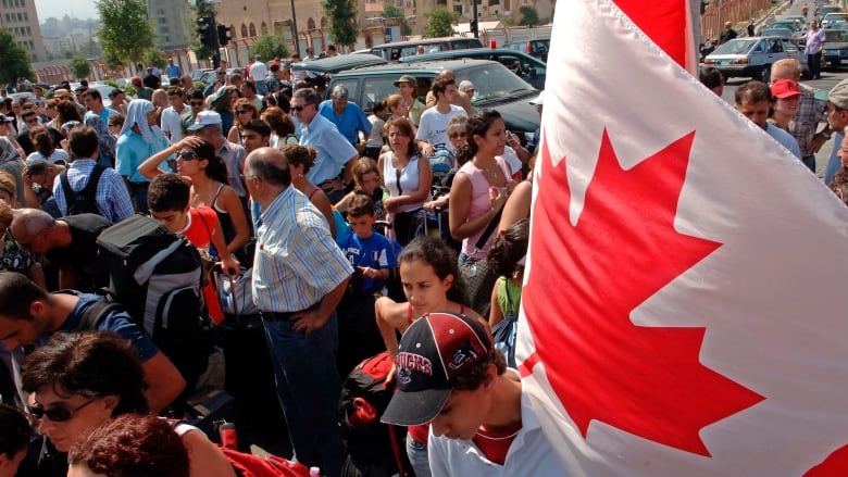 A Canadian flag is held up, on the right side of the image, above a large crowd of people stand outside.