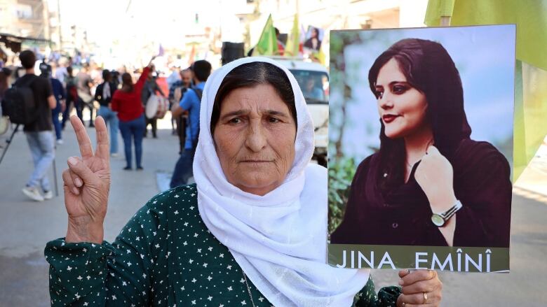 An older woman in a headscarf holds up a sign bearing the picture of a younger woman with the inscription on it, 'Jana Amini.'