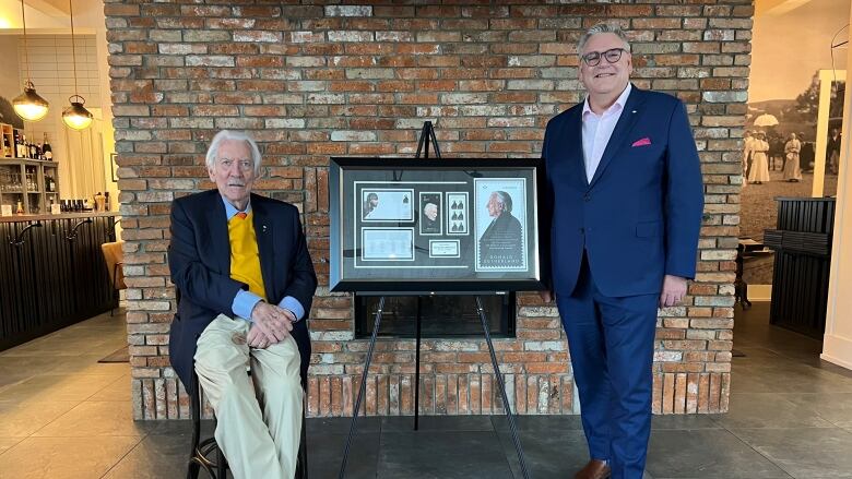 Two men, one elderly and seated, are shown posing for a photo on either side of a black frame.