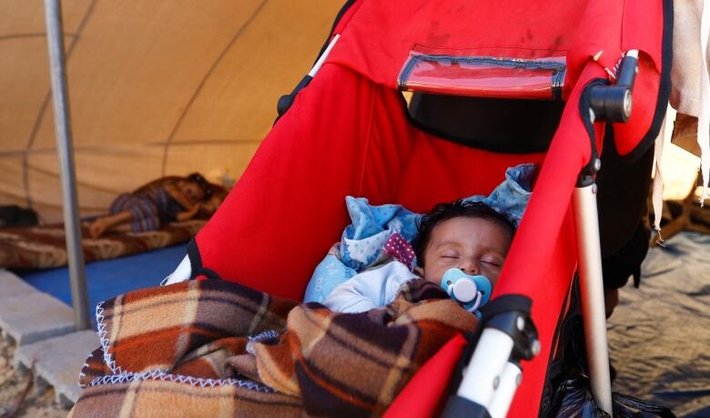 A baby with a blue soother and black hair sleeps in a red stroller inside a humanitarian aid tent.