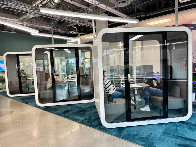 A row of three large glass boxes, like phone booths, big enough for two people to sit in, in a bright office space.