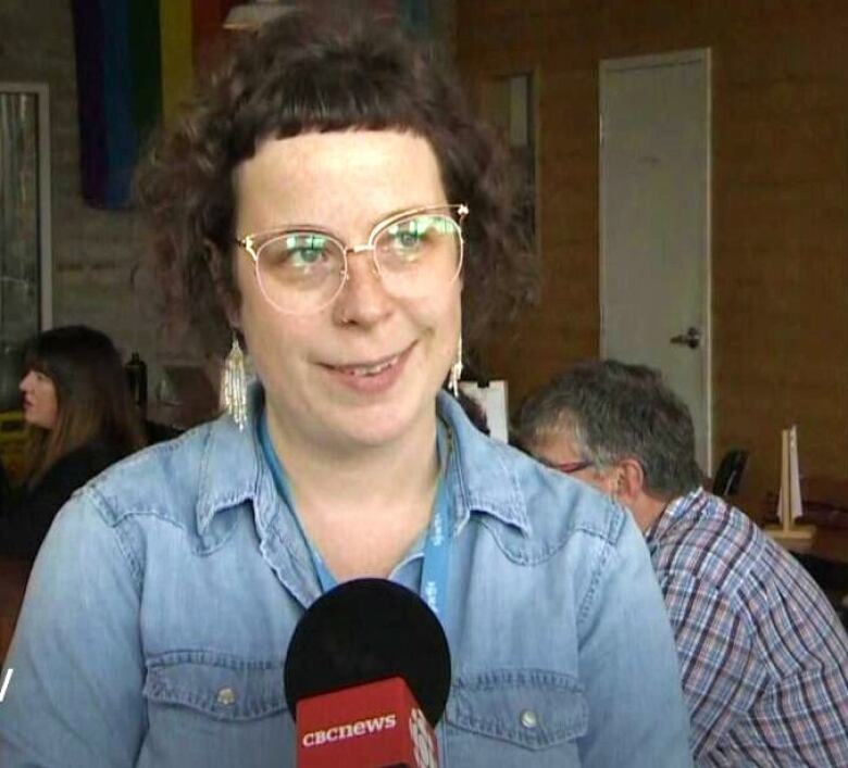 A woman with short hair, glasses, and a denim shirt stands in front of a microphone.