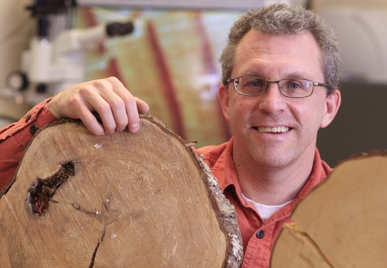 A man in an orange shirt stands with his hand resting on a large piece of cut wood.