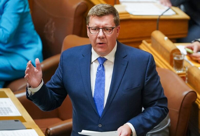 A man in a blue suit stands and speaks in a legsislative chamber.