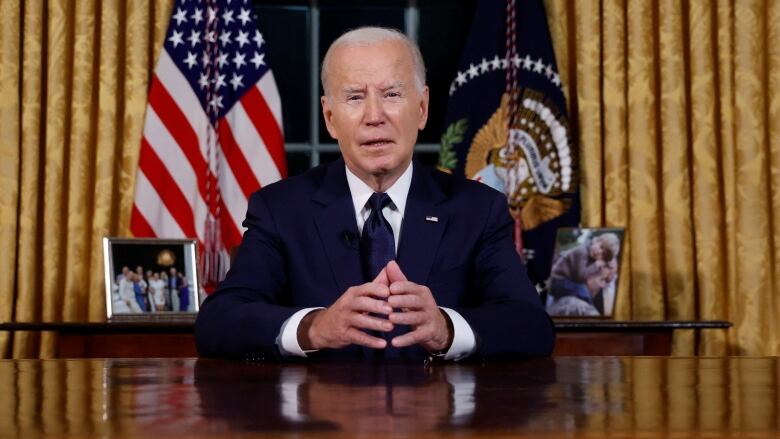 White haired man in blue suit sitting at des speakign to camera