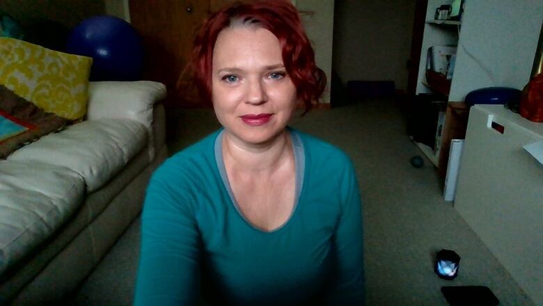 A red-headed woman in a teal shirt sits in a carpeted room with white couches