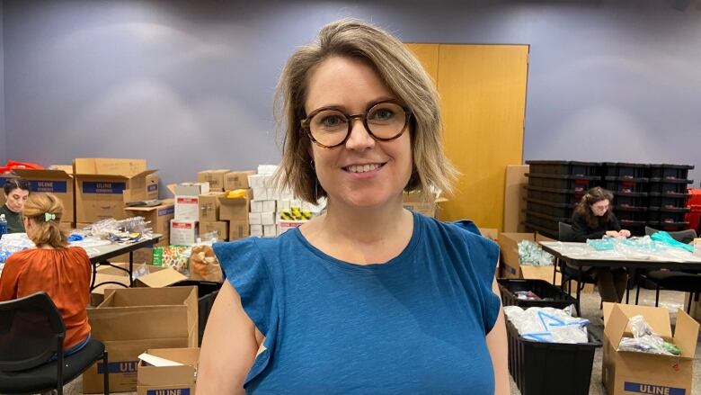 A woman wearing glasses and a blue top smiles at the camera. Open boxes and people working at tables can be seen behind her.