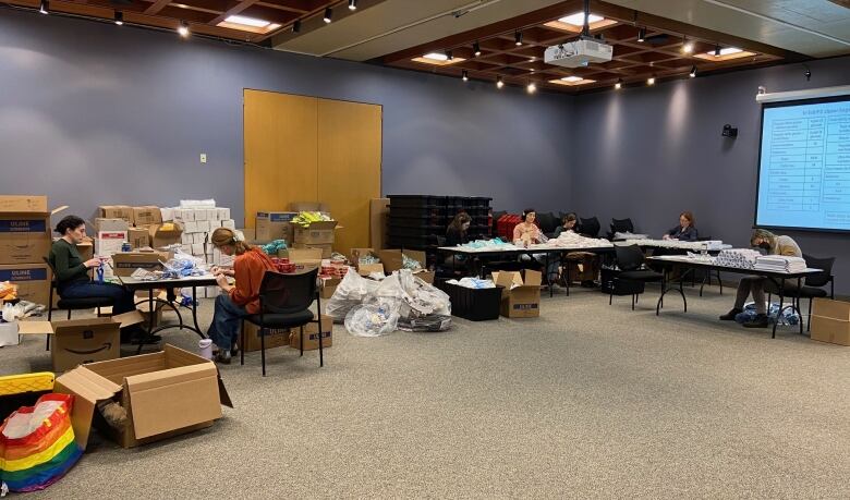 A large room with several tables in the background. People sitting at those tables are surrounded by open boxes and supplies.