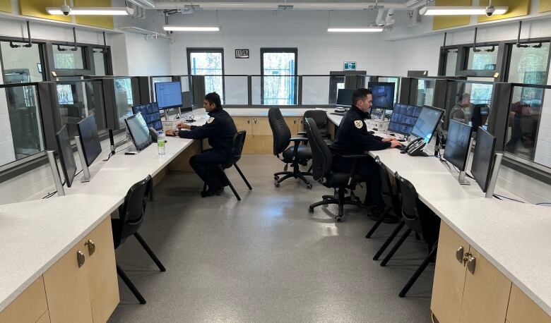 Two male guards sit at desks in a plexiglass-surrounded office pod.