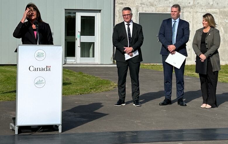 A woman speaks at a podium while two men and a woman in business suits stand nearby.