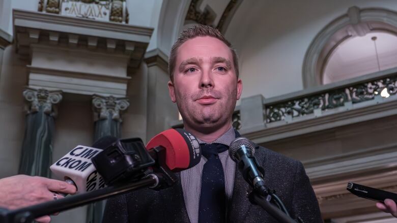 A man in a grey suit and blue tie speaks at a microphone.