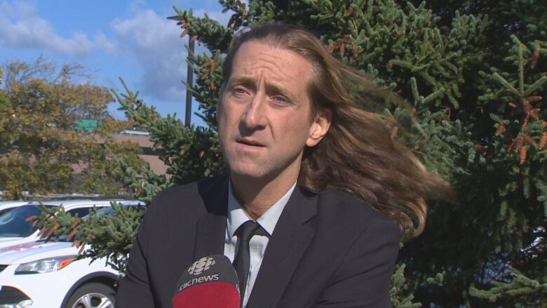 A man with long brown hair wearing a suit sits outside in front of a microphone.