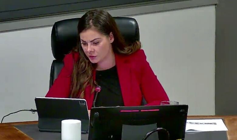 A city councilor sits behind a computer monitor and reads from the screen of a tablet.