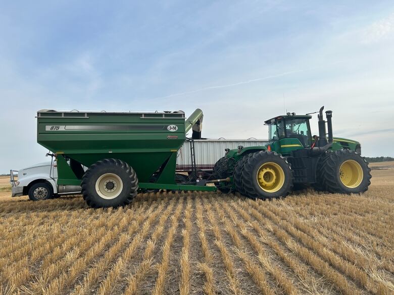 A green tractor and green grain cart are pictured in a field of stubble loading a canola crop into a semi trailer.