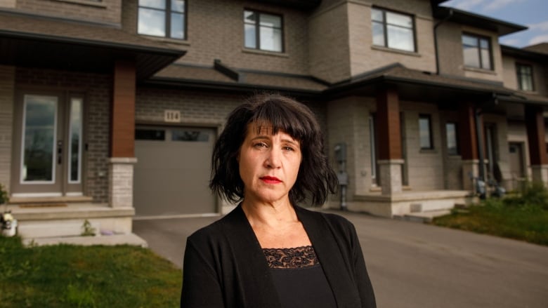 A white woman in a black shirt and cardigan stands in front of a grey-bricked townhouse. 