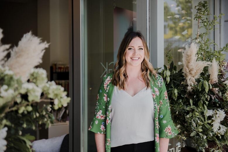 A smiling white woman with curled blonde hair wearing a green silk and floral shirt over a white t-shirt stands in front of a store, flanked by plants. 