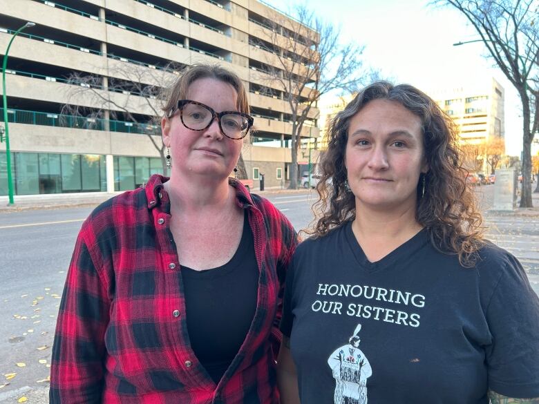 Two women stand together on a sidewalk, facing the camera.