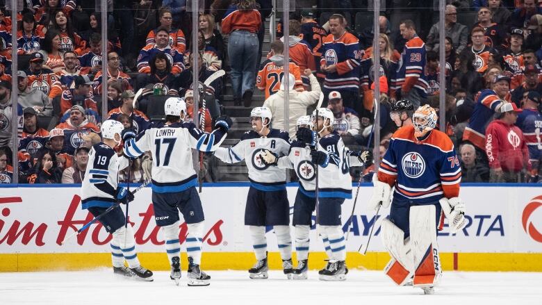 Players in white jerseys gather on an ice rink next to a goalie in a blue jersey.