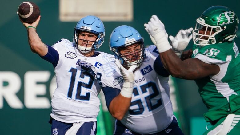 Toronto Argonauts quarterback Chad Kelly is throwing a football while a defender is shoving a Regina Roughrider away