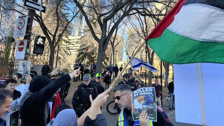 A police officer talks to protesters waving a Palestinian flag on a street next to a park.