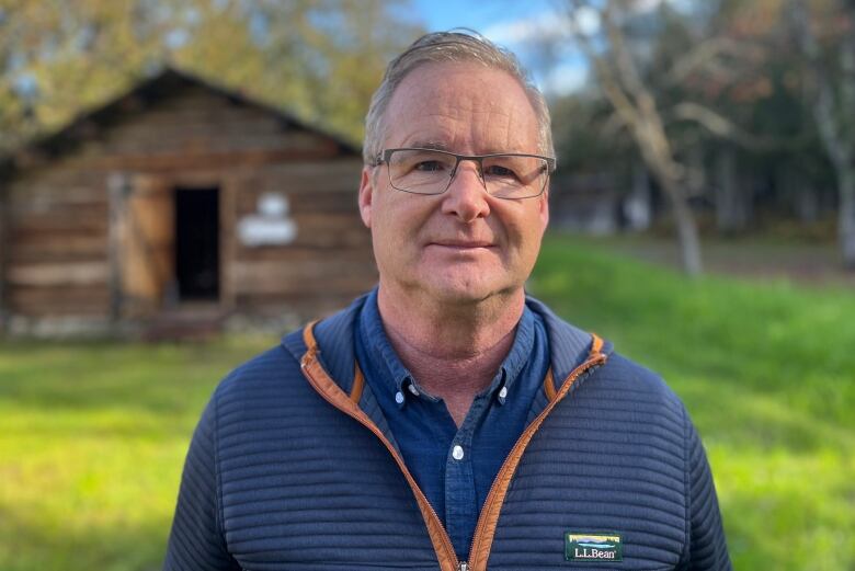 An older man with glasses standing outside in front of a wooden cabin. He is wearing a blue shirt and blue L.L. Bean jacket overtop.