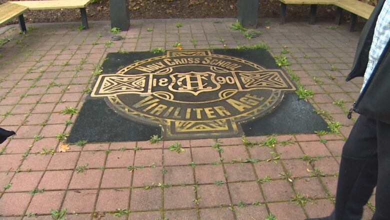 A photo of a plaque planted in the ground, surrounded by walkway pavers. There's an unidentified man to the right. 