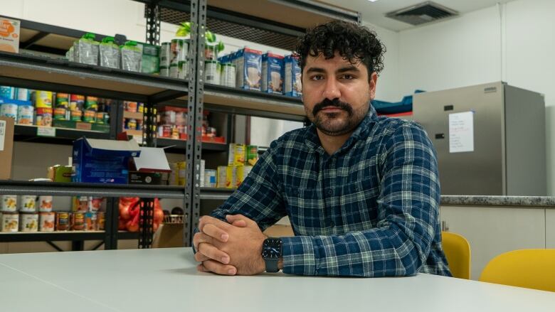 A young man sits at a table with his hands folded. Behind him are shelves filled with food.