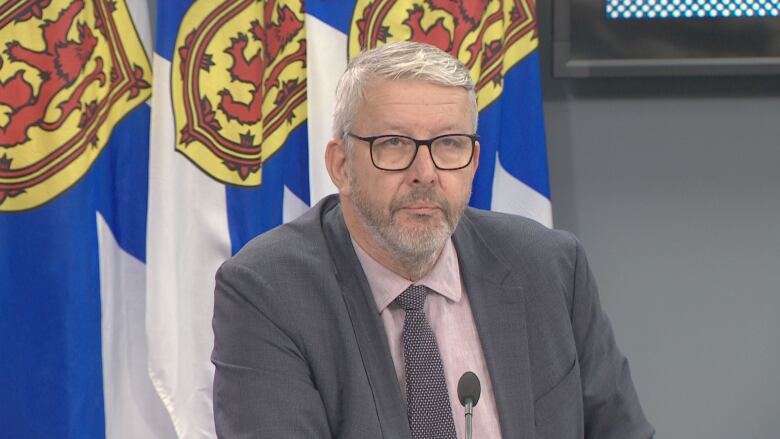 A white-haired man with glasses, who is wearing a blazer and a pink oxford shirt with a grey tie, speaks to reporters.