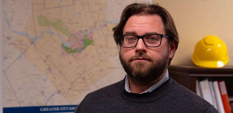 A man with a beard stands in front of a map with yellow hardhat beside him