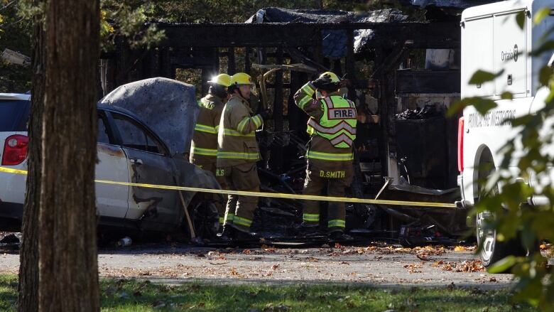 Three firefighters can be seen standing in front of a charred wooden structure. One holds up a piece of scorched metal. A white vehicle can be seen next to them, it's front end burned.