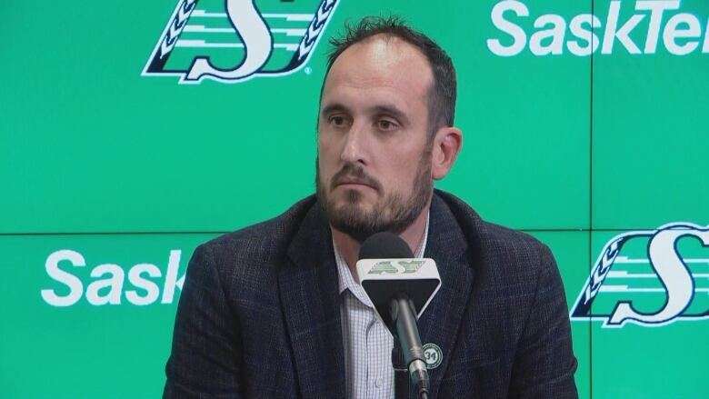 A man in a suit speaks at a microphone. Behind him is a green backdrop with the Saskatchewan Roughriders logo.