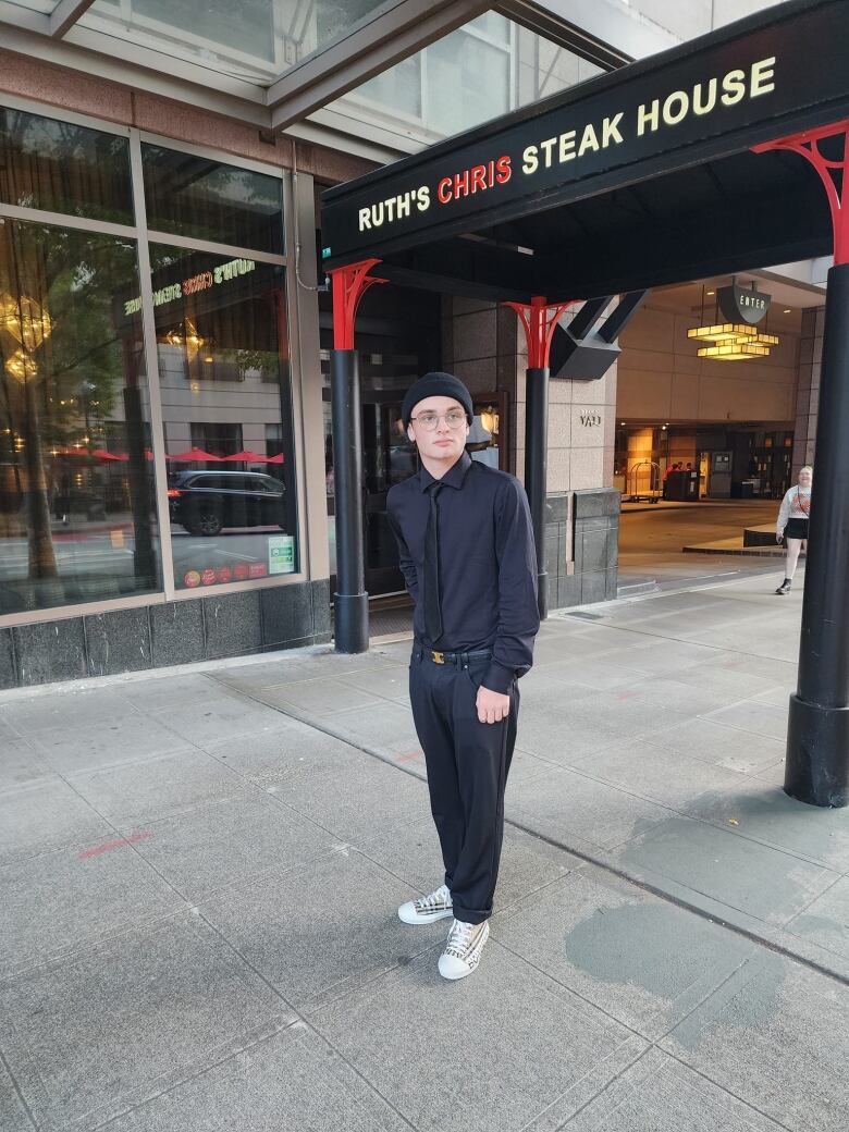 A teenager wearing a black suit poses in front of a restaurant.