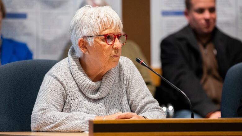 An old woman with red glasses sits pursing her lips in front of a mic.