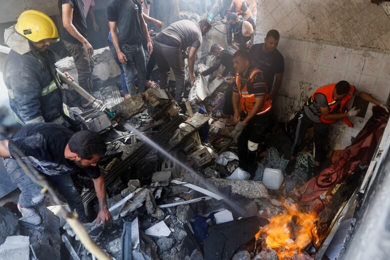 Several people including first responders in vests are shown in a tight space inside a home filled with debris and damage. One person sprays water at a fire in the corner.