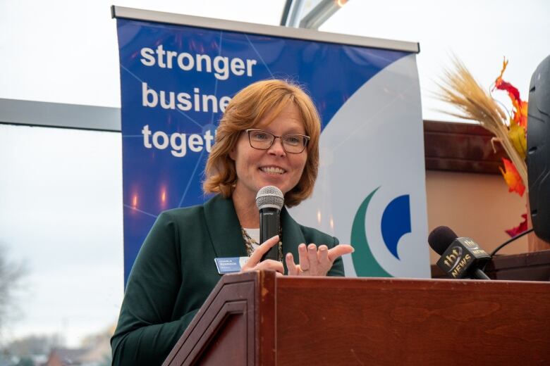 A woman with red hair and glasses speaks into a microphone at a podium.