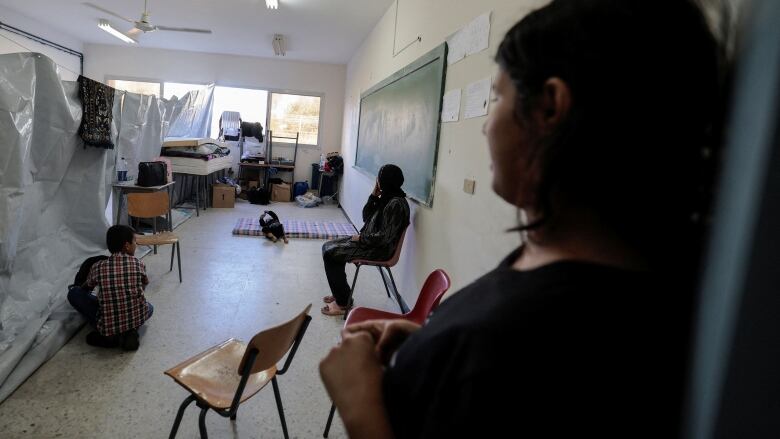 Three people sit and stand amid some chairs and a mattress in a classroom that has been repurposed as a refugee shelter.