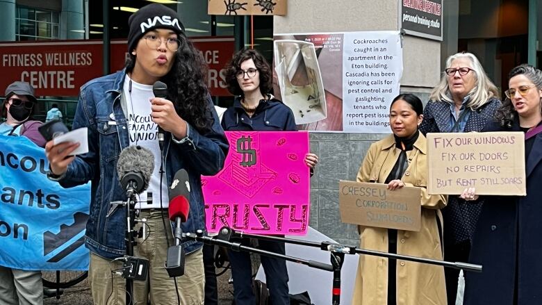 A person speaks on a mic as others hold up signs denouncing Cressey Corporation as slumlords and one reading 'Fix our windows, fix our doors, no repairs but rent still soars'.