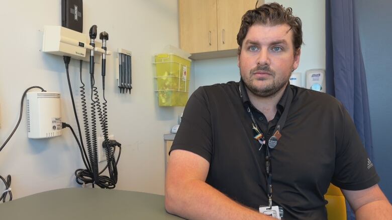 A doctor in a black short sleeved collared shirt is shown in an examination room with a naloxone kit on the wall behind him.