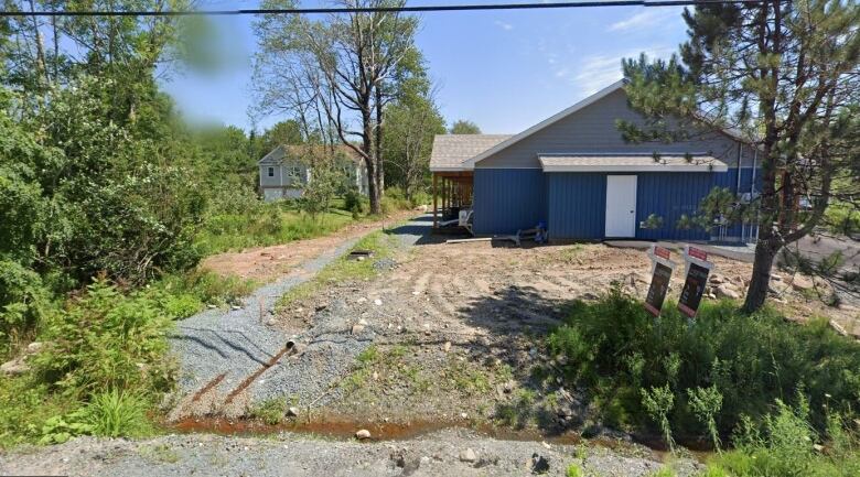 There are some scattered trees and brush along the property line, but the home on the left is much more open to the street and neighbouring property. A flattened gravel area is between the properties, to the left of a one-story blue building