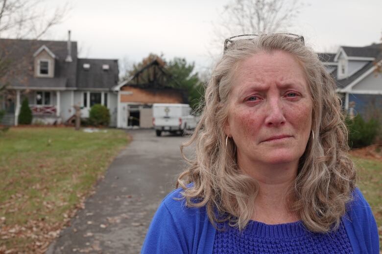 A woman with curly grey-blonde hair, her eyes red from crying, stares at the camera with a sad look on her face. Behind her is the burned remnants of a garage.